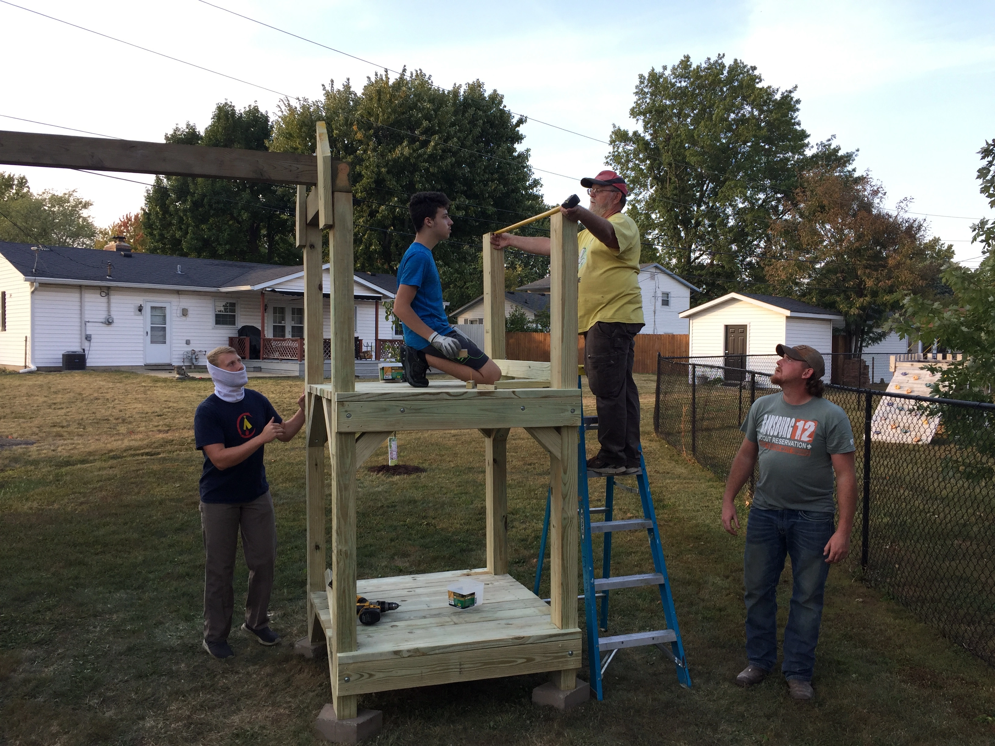 Two scouts out of uniform and a volunteer observing another volunteer as he measures the distance between two posts on a piece of playground equipment that is currently being worked on