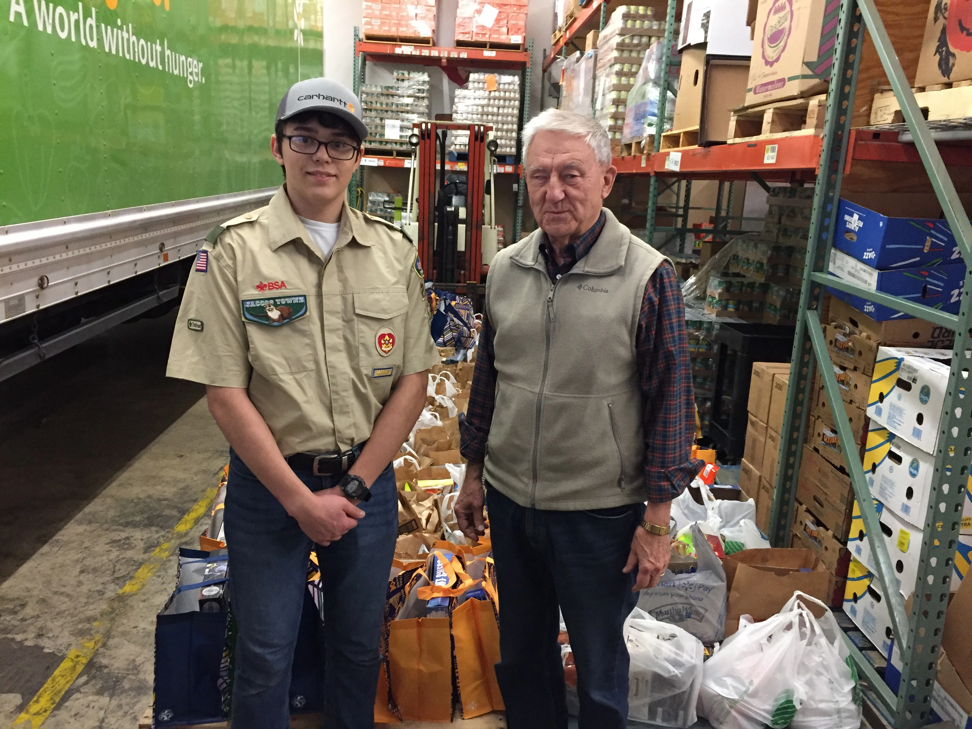 Alejandro Garnica and a volunteer standing together infront of lunch bags created for Eagle Scout project inside of a warehouse